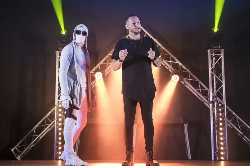 R'Key et son personnage masqué en combinaison blanche, entre deux structures lumineuses, sous les projecteurs