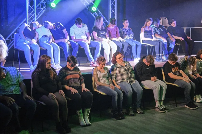 Deux rangées de volontaires assis, tête baissée, plongés en hypnose sous les projecteurs